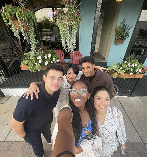 A group of 5 Cencora interns smile and pose together as they take a selfie. They stand in front of a restaurant that has blue exterior walls, window boxes, and hanging plants with vines.