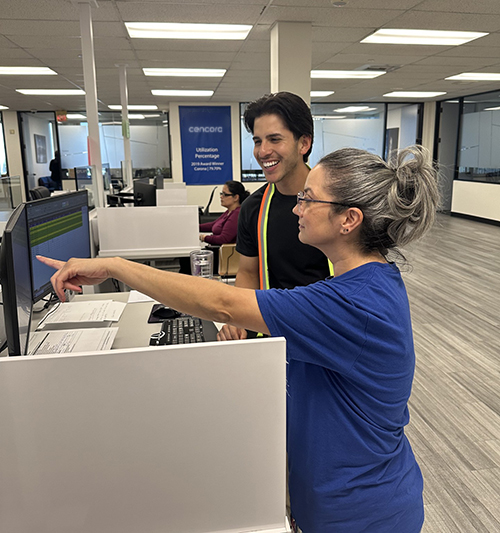 An intern works with a team member. The intern is smiling as the team member is showing them something on their computer. They are at a standing desk.