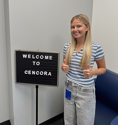 A Cencora intern smiles at the camera and gives two thumbs up. They are standing next to a sign that says "Welcome to Cencora".