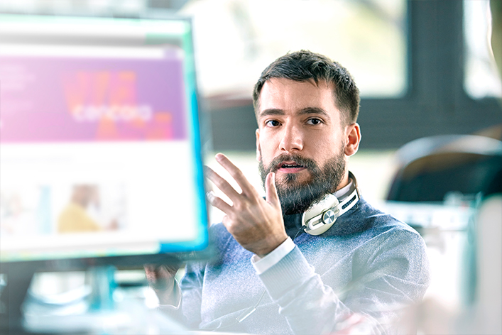 A light-skinned office worker wearing headphones around their neck explaining something with hand gestures from behind a desktop computer monitor.