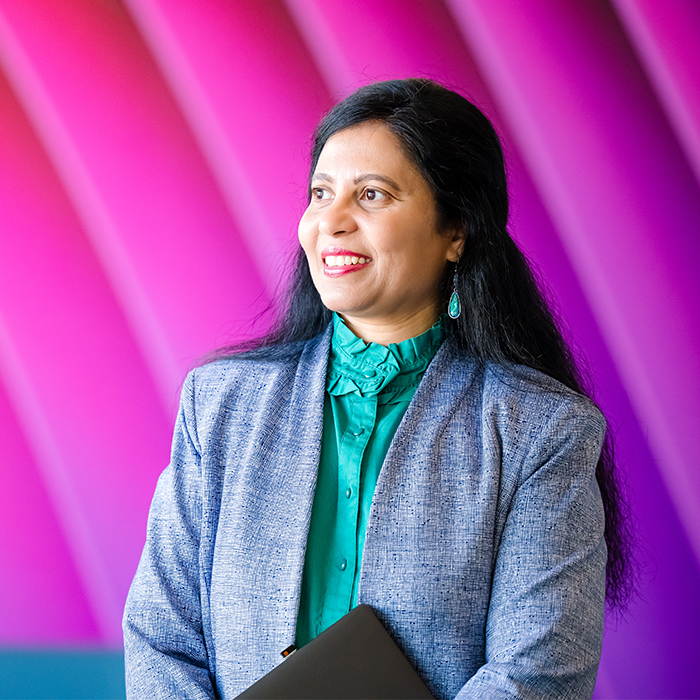 A Cencora team member stands in front of a colorful magenta wall. They smile and look off to their left - they are wearing business professional clothing - a green top and a grey business jacket.