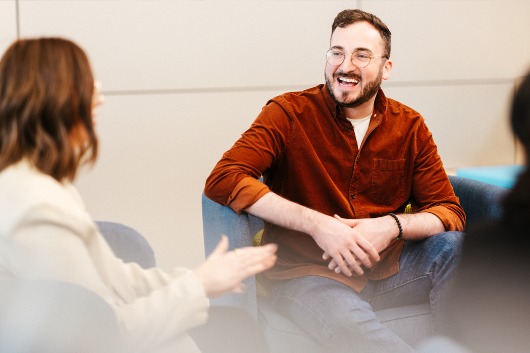 A Cencora team member in a red shirt and glasses is smiling and conversing with another person in a white jacket while sitting on blue chairs.