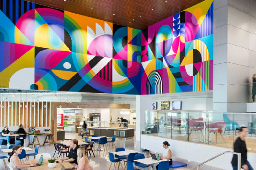 Inside the Cencora headquarters, a wide angle and distanced view of several people sit at tables and chairs, conversing, in an open contemporary office space where a large, vibrant abstract mural adorns the walls.