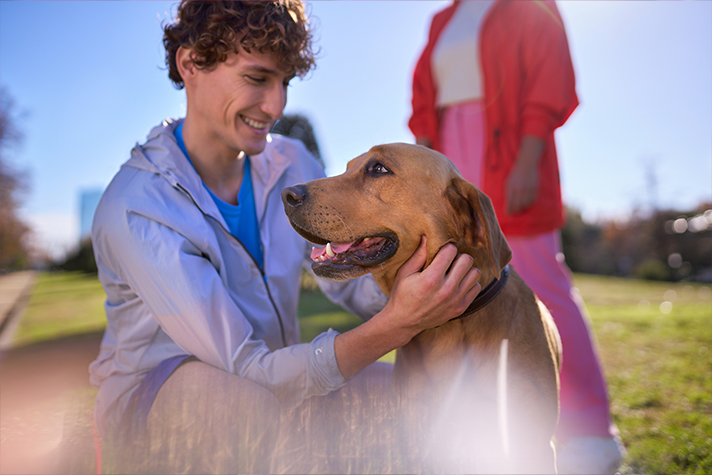 A light-skinned person in a white track jacket kneeling down to pet a dog in an outdoor park setting during a sunny day.