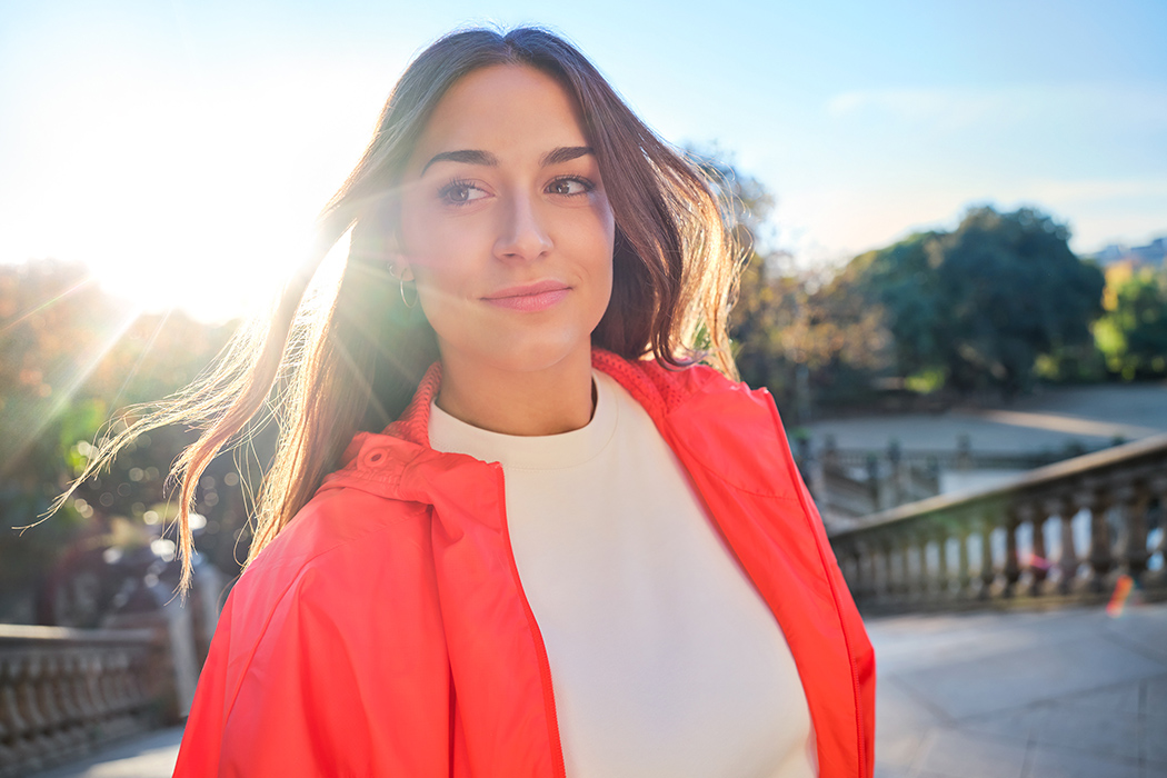 A person stands outside and smiles confidently while looking off camera. The sun is setting and shining behind them.