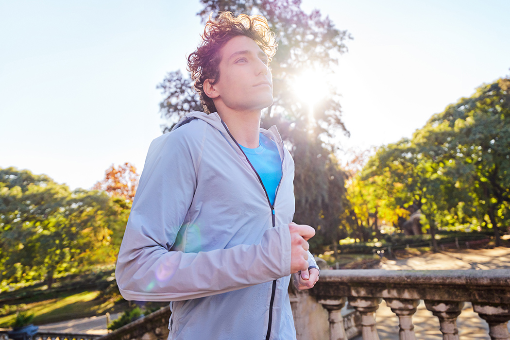 A light-skinned person wearing a white track jacket and a bright t-shirt jogging through a park with the morning sun shining behind their head.