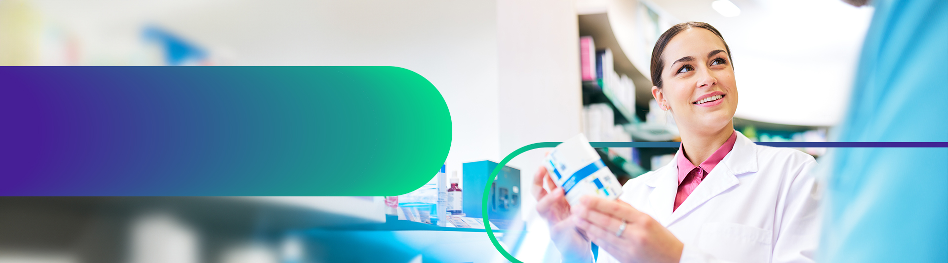 A pharmacist holds up a box of medication to show another person who is mostly out of frame. They wear a pink collared shirt beneath a white lab coat. Shelves of medication are located behind them.