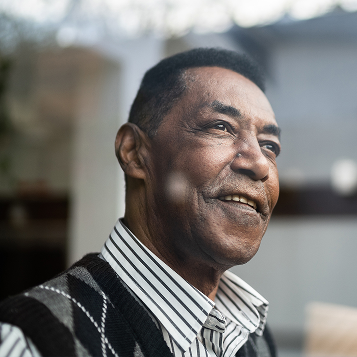 A senior man dressed in a collared shirt and a sweater vest looks out a window with a pensive look on his face. 