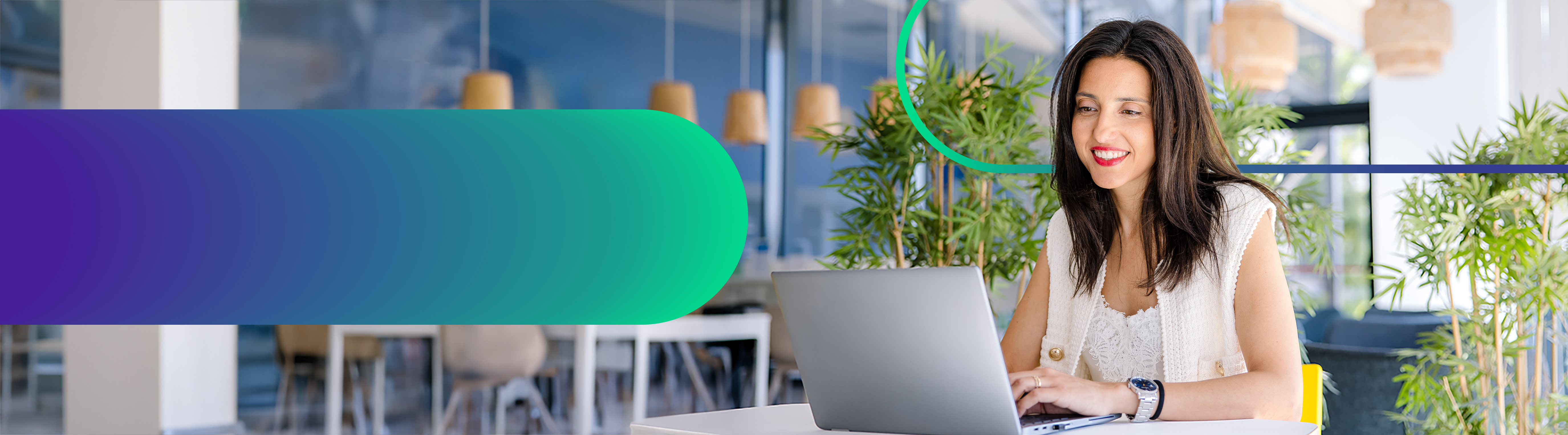 A Cencora team member sits in a naturally lit shared space in our Spain office while working on a computer. In the background, there are plants and rattan lights.