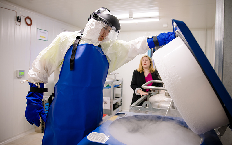 A Cencora team member opens a cold storage unit - they are wearing a protective suit with gloves, an apron, and face shield. 