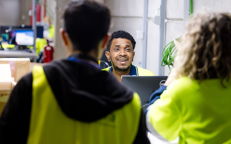 3 Cencora team members in high visibility vests are engaged in conversation. The one team member is at their computer and smiling while looking across the table to the other two.