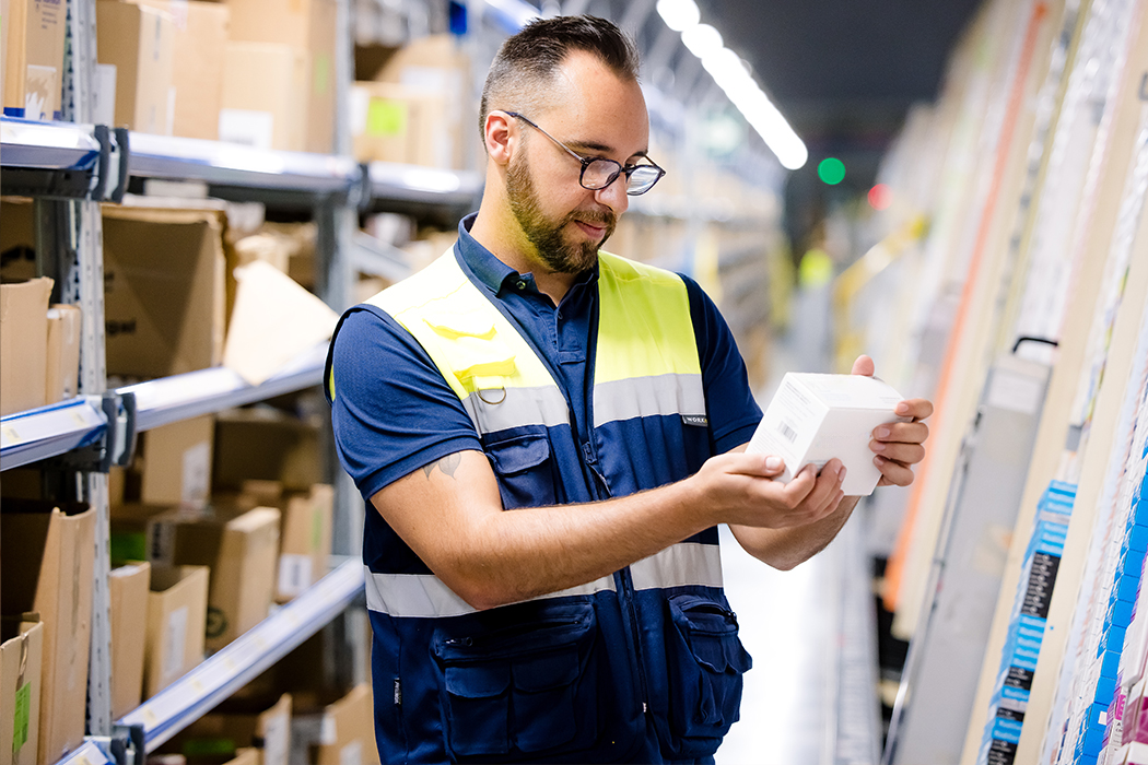 A Cencora team member wearing glasses and a high visibility vest stands in a facility holding a product box and inspecting it.