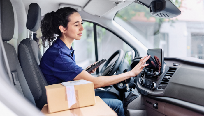 A delivery driver types an address into the dashboard screen of her delivery van. Cardboard boxes are stacked up on the passenger seat.