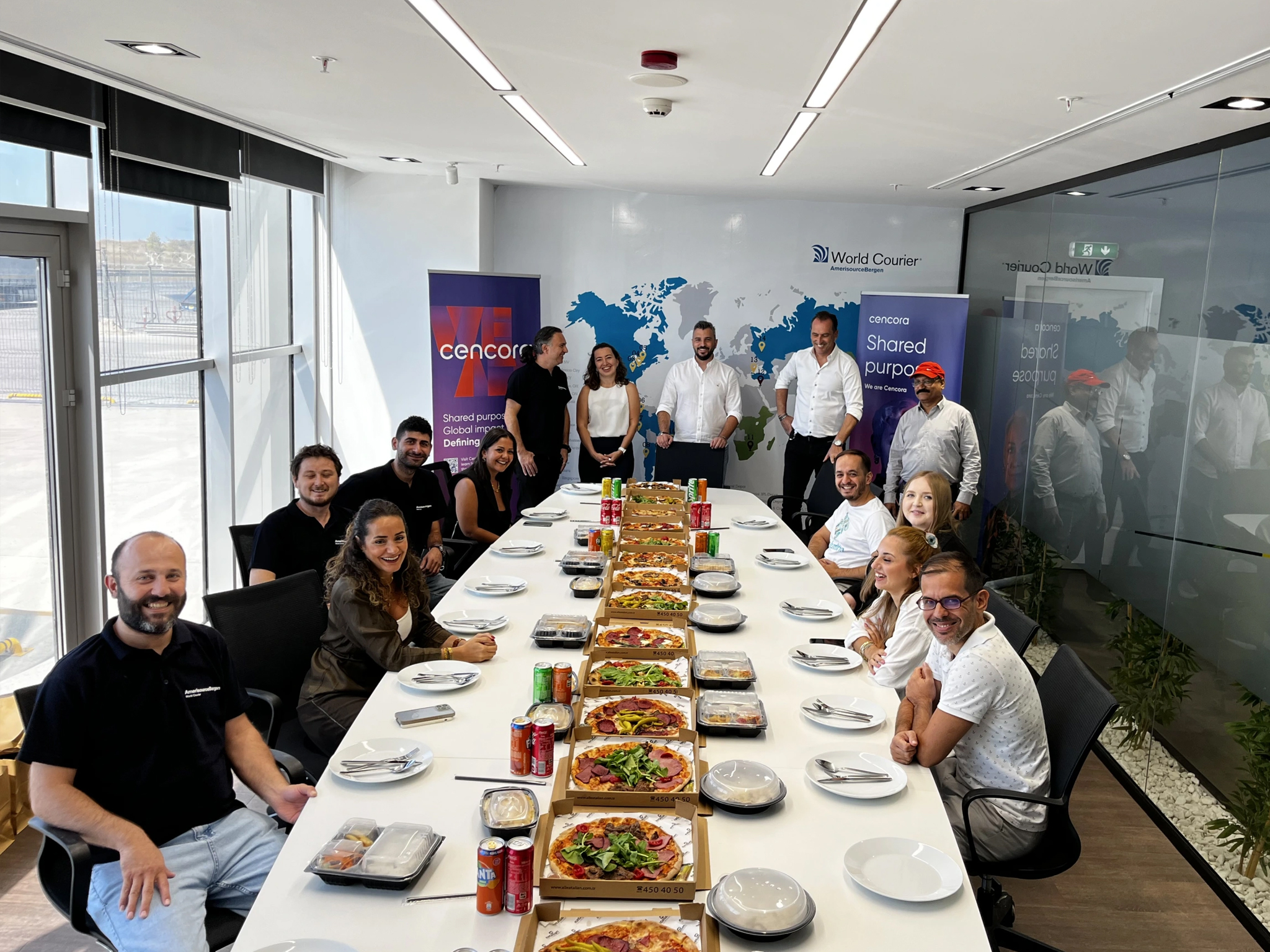 A group of Cencora team members pose and smile for the camera at a conference table that has pizzas down the center.