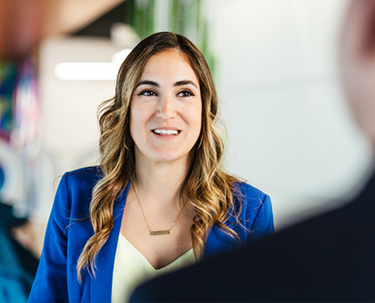 A Cencora team member with long curly hair wearing a blue blazer smiles while talking to another team member standing opposite. They are standing in the hall of the Cencora headquarters building.