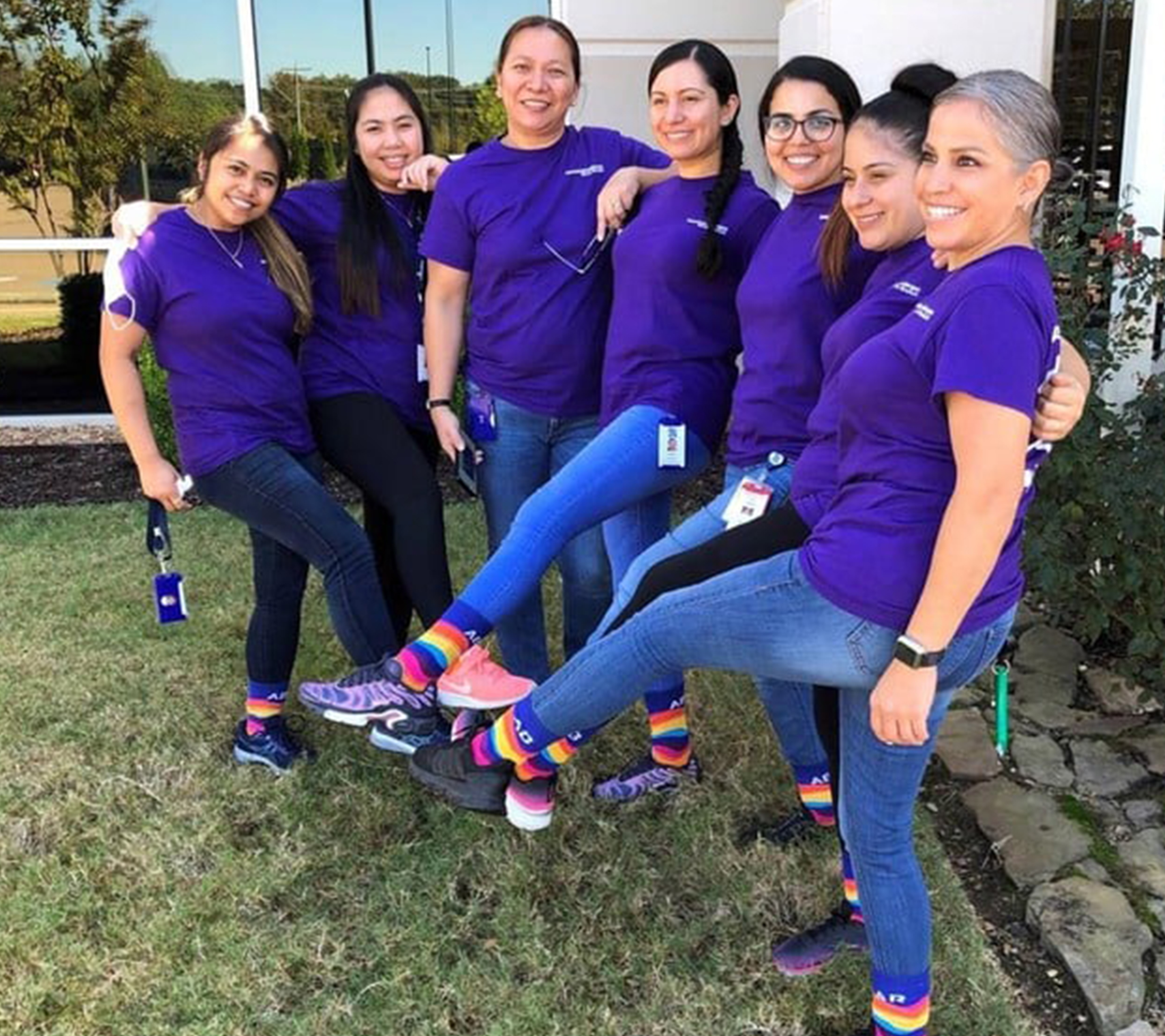 A group of WIN ERG members stand together with their arms around each other smiling for a photo. They are showing off their rainbow colored Cencora socks.