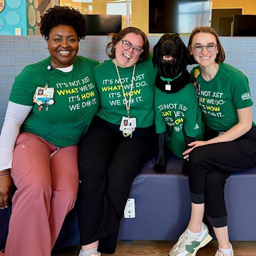 A group of smiling child life specialists sit with a facility dog at Children’s. 