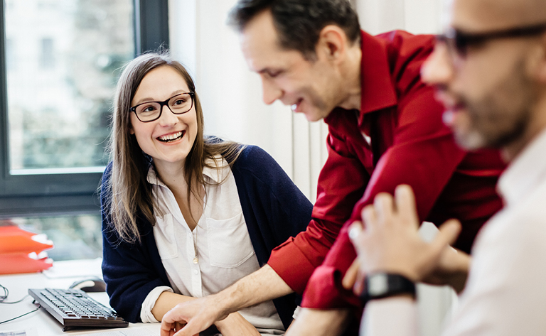 Two employees standing in an office side by side with group of employees working together behind them.