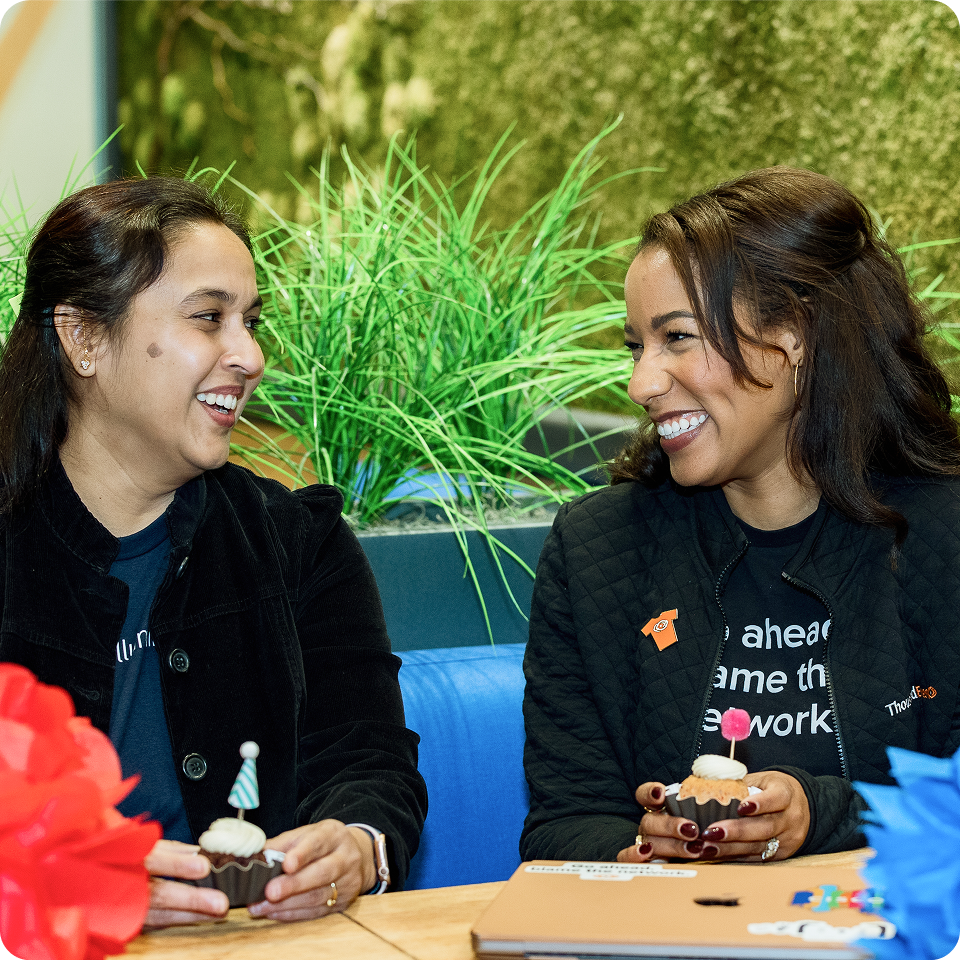 Two smiling women look at each other as they both hold cupcakes.