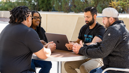  Four people sit outside around a table with one wearing a We Are Cisco t-shirt looking at a laptop.