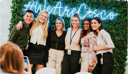 A group of young Cisconians smiling in front of a green wall with a neon #WeAre Cisco sign.