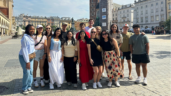 A group of about thirteen young people standing together and smiling in a large European city square, with historic buildings and a tall stone tower visible in the background.