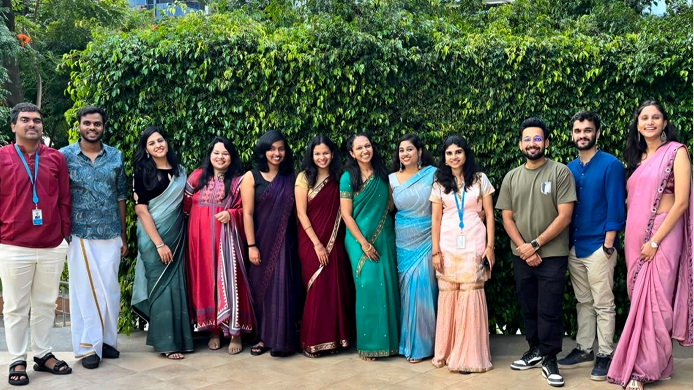 A group of twelve people standing in a row and smiling for a photo in front of a tall green hedge.