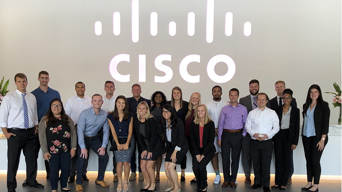 A group of about eighteen professionals in business attire posing for a photo indoors in front of a large wall bearing the Cisco logo.