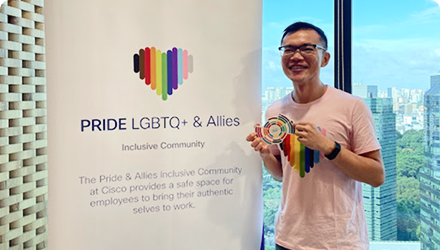 Justin stands in front  of an office window and a Pride LGBTQ+ & Allies banner.
