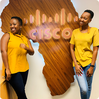 Two black women smile in front of a wooden map of Africa with the Cisco logo on it.