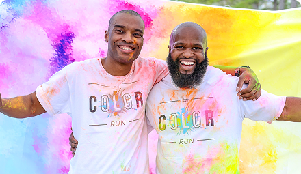 After finishing a color run, two men stand in front of a colorful backdrop smiling with bursts of color sprinkled all over their shirts and bodies.