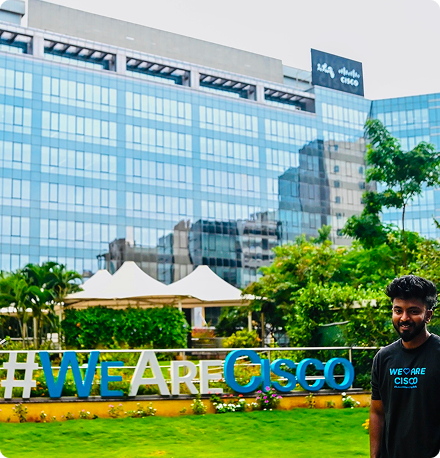 A smiling young man stands in front of a Cisco office building and a #WeAreCIsco sign wearing a We Are Cisco t-shirt.