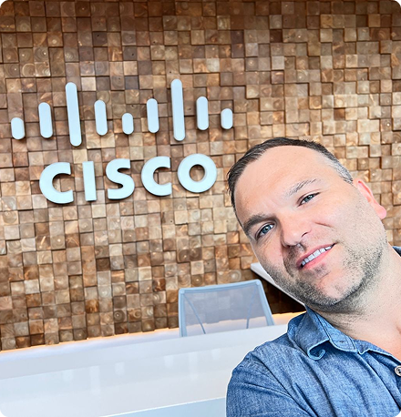 A man smiles while taking a selfie in front of a wood block wall with a Cisco sign in one of our offices.