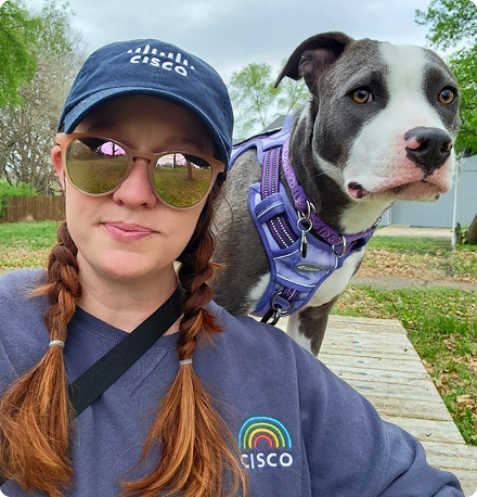 A woman decked out in Cisco hat and sweatshirt looks at the camera with her dog peering over her shoulder.