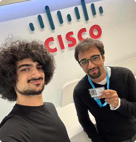 Two young men stand in front of a Cisco sign in an office. One holds his badge up to the camera.