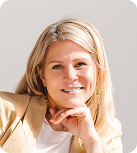 A young blonde woman with long hair smiles with her chin on her hand while wearing a white shirt under a beige blazer.
