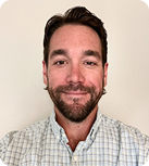A dark-haired man with a beard wearing a white, green, and and brown checked button-down shirt stands in front of a white wall and looks thoughtfully at the camera.