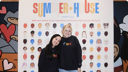 Two women stand in front of a backdrop with caricatures of human faces. The backdrop says "Summer House" at the top.