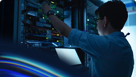 An IT professional stands in front of a server making adjustments while looking at a laptop.