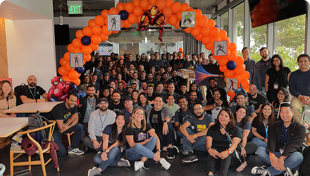 A large group of Thousand Eyes employees sits under an orange balloon arch with pictures of superheroes like Ironman, Captain America, Captain Marvel, the Hulk and Spiderman  on it.