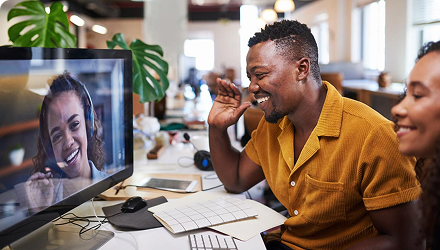 A man sits at a desk in an office waving to a colleague on a screen in front of him.