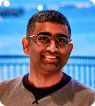 A young East Asian man wearing glasses and a brown henley with a black color smiles in front of a blue background with a blurred metal fence.