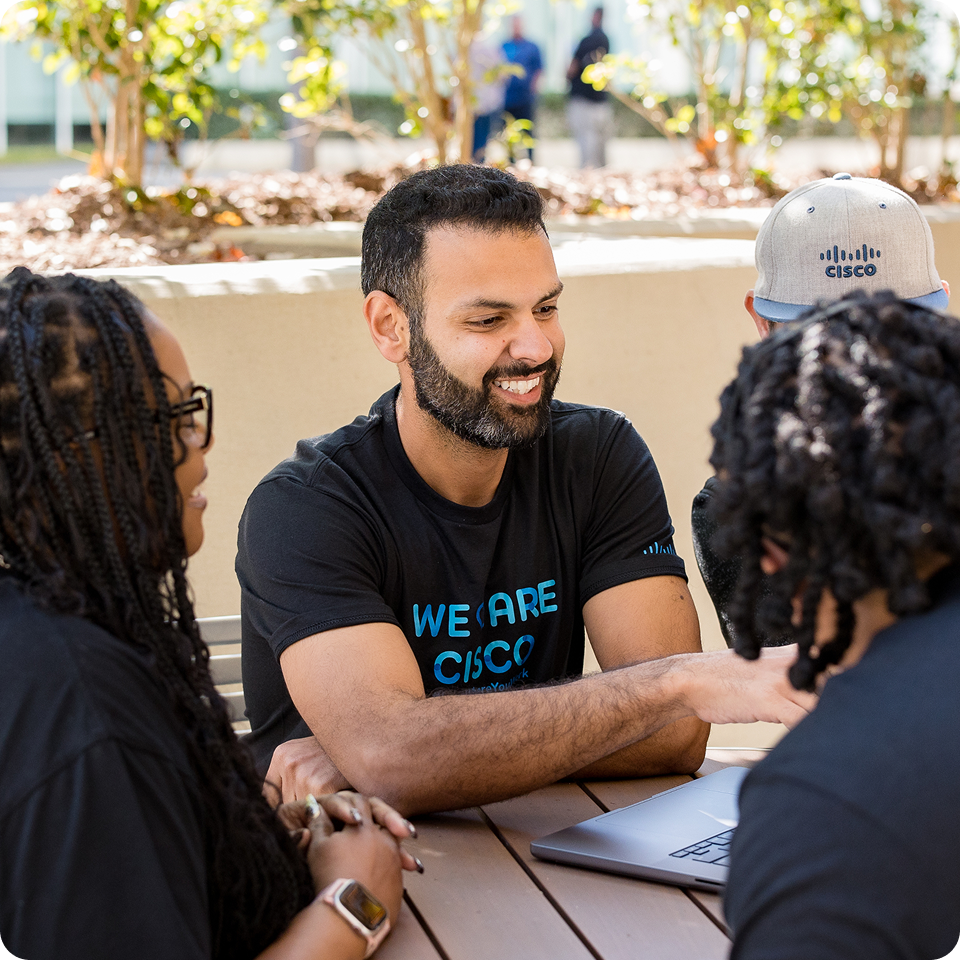 A smiling man wears a "We Are Cisco" t-shirt in conversation with a table of people.