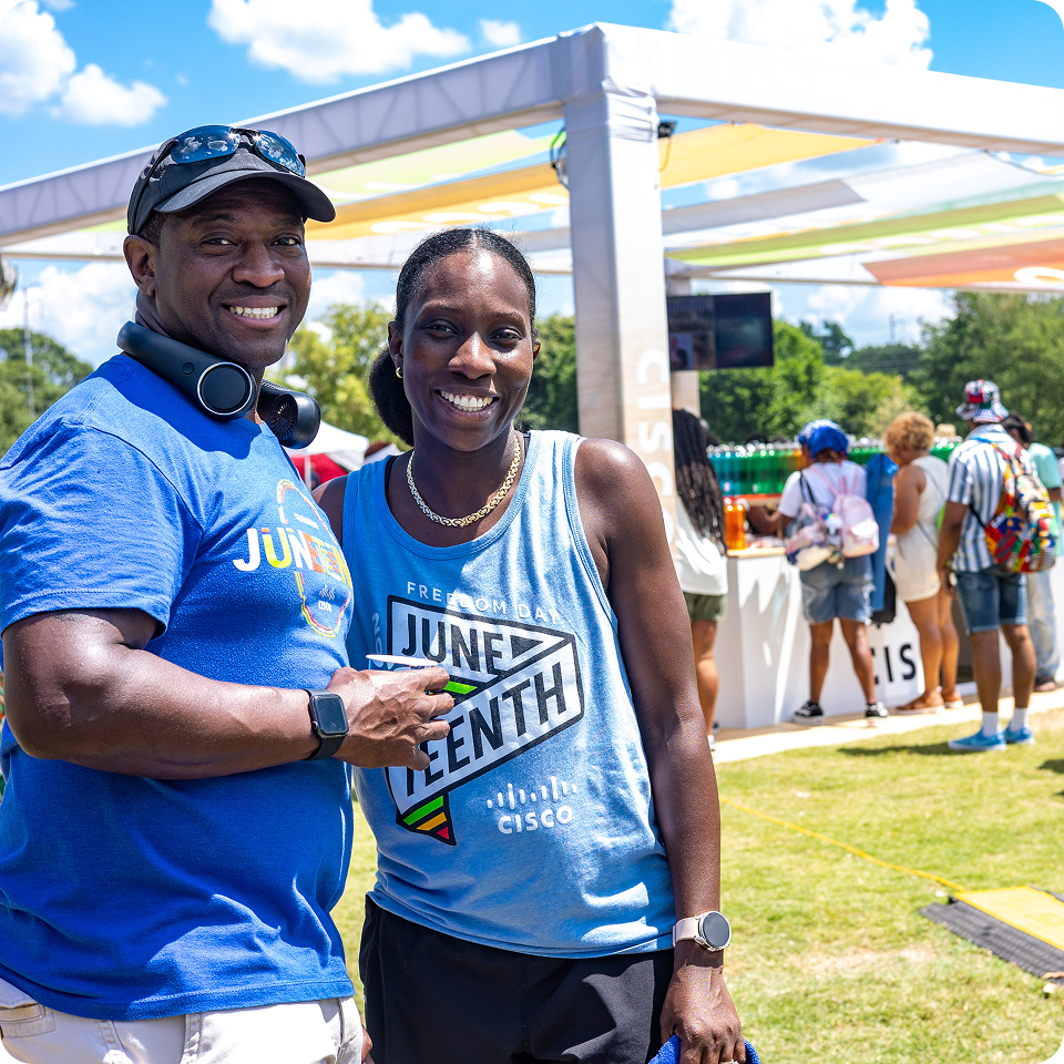 An outdoor Juneteenth celebration with two Black individuals in the foreground smiling at the camera.