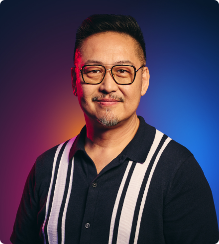 An Asian man wearing glasses and a blue casual button-down shirt with shite stripes on either side looks thoughtfully at the camera.