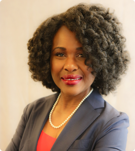 A Black woman wearing a coral blouse under a charcoal gray suit jacket with pearls and bright lipstick smiles at the camera.