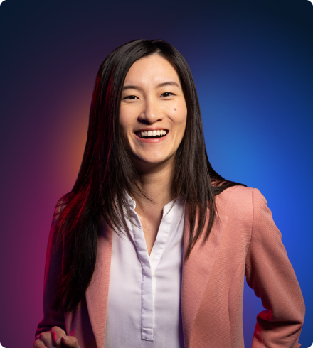 An Asian woman wearing a white blouse under a pink blazer looks like she's laughing with a big smile on her face.