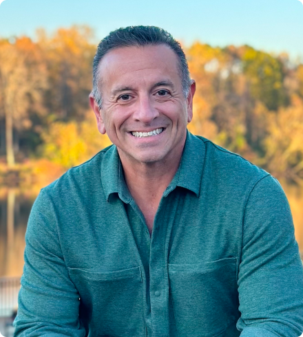 A man wearing a casual green button-down shirt sits in front of a blurred stand of trees in autumn colors smiling at the camera.