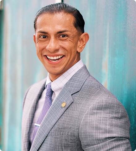 A Latino man wearing a gray patterned suit jacket with a gold lapel pin over a light-purple checked shirt with a purple patterned tie gives a beaming smile at the camera.