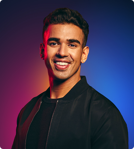 A young man wearing a black t-shirt with a black zip-up bomber jacket over it smiles confidently at the camera.